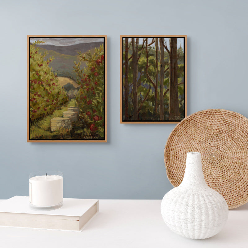 Two framed oil paintings of Tasmanian landscapes on a wall with a vase, candle, book, and woven tray on a table below.