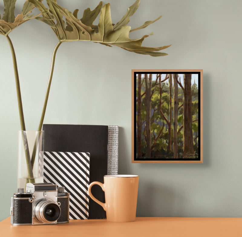 Decorative setup with a framed oil painting of a Tasmanian eucalyptus forest, camera, mug, and books on a table against a light green wall.